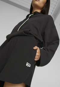Svart hettegenser med snor, kombinert med svarte shorts med et hvitt Puma-logo og sideposettdetaljer.