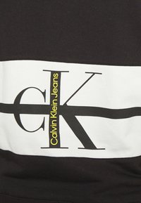 Schwarzes Sweatshirt mit einem weißen horizontalen Streifen, mit großem schwarzen "CK"-Logo und dem Text "Calvin Klein Jeans" in Gelb. Weicher Baumwollstoff.