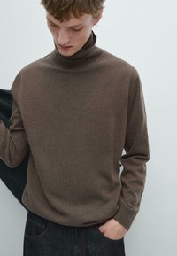 Brązowy sweter z golfem wykonany z miękkiego materiału, charakteryzujący się luźnym krojem i długimi rękawami, połączony z ciemnymi dżinsami oraz gładkim tłem.