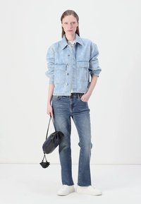 Veste en denim bleu clair avec deux poches avant, accompagnée d'un jean bleu moyen et de baskets blanches. Le modèle tient un sac noir.