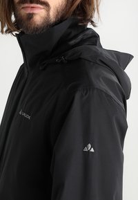 Homme avec barbe portant une veste imperméable noire avec capuche et logo Vaude sur la poitrine et la manche, face légèrement tournée à gauche.