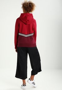 Röd hoodie med en vit och svart randig detalj på baksidan, kombinerad med svarta plisserade vida byxor och vita sneakers.