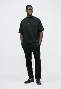 Μαύρο βαμβακερό T-shirt με κοντά μανίκια, με λευκή επωνυμία. Συνδυάζεται με μαύρα παντελόνια joggers και μαύρα γυαλιστερά παπούτσια. Απλός σχεδιασμός.