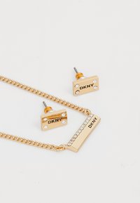 Set di gioielli DKNY color oro composto da una collana con catena e pendente rettangolare, accompagnato da orecchini a perno impreziositi da strass.