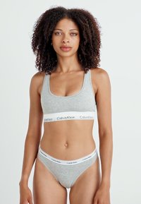 Szürke pamut bralette és tanga szett, széles fehér pánttal, amin "Calvin Klein" logó található. Egyszerű, testhezálló kialakítás, minta nélkül.