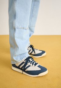 Marineblaue und graue Sportsneaker mit Schnürsenkeln, ausgestattet mit einem strukturierten Obermaterial, durchbrochenem Design und beiger Sohle, kombiniert mit hellblauen Denim-Jeans.