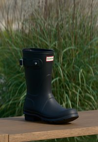 Čierna gumová dažďová obuv s okrúhlou špičkou, detailom pracky a bielym logom "HUNTER". Lesklý povrch, robustná podrážka, umiestnená na drevenom povrchu.