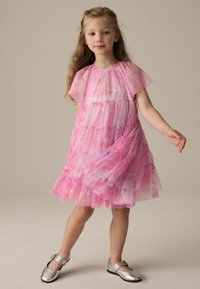 Mehrlagiges pinkes Kleid mit durchsichtiger, geraffter Oberseite und wellenförmigem Muster. Kombiniert mit glänzenden silbernen Schuhen mit einer Schnallen-Details.
