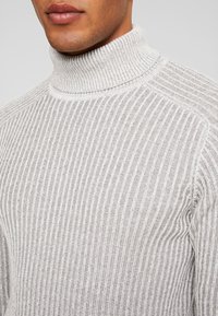 Grauer, gerippter Rollkragenpullover mit hohem Kragen und strukturiertem Stoff. Die vertikalen Streifen verleihen visuelles Interesse und Tiefe.