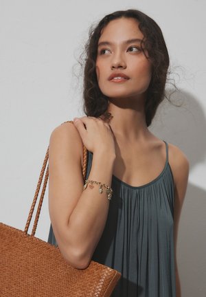 Junge Frau mit lockigem Haar in einem ärmellosen grauen Kleid, die eine geflochtene braune Schultertasche hält und ein Charm-Armband am linken Handgelenk trägt.