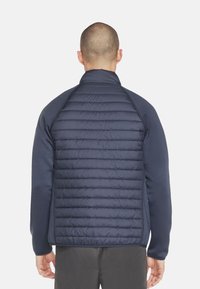 Marineblauwe jas met een gevoerde achterzijde van pufferstof en solide panelen op de mouwen. Hoge kraag en elastische manchetten. Glad textuur en lichtgewicht ontwerp.