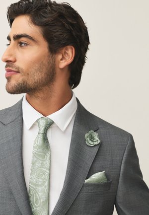 Man in gray suit with white shirt, light green paisley tie, matching pocket square, and floral lapel pin, facing left against plain background.