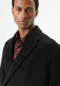 Zwarte wollen jas met een reverskraag, gestructureerde stof en een licht bordeauxrood overhemd eronder met bijpassende knopen.