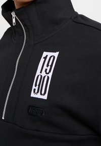 Czarna kurtka zapinana na zamek z wysokim kołnierzem, z białą grafiką "1990" i teksturowanym logo naszywką na dolnej części przodu. Materiał mieszany bawełniany.