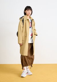 Manteau imperméable beige avec un col surdimensionné, grandes poches, porté sur un T-shirt graphique blanc, associé à un pantalon marron et des baskets multicolores.