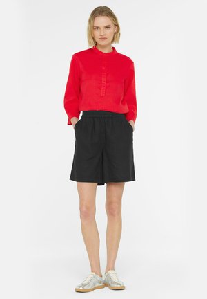 Rotes Langarm-Hemd mit Knopf und Stehkragen, kombiniert mit schwarzen, lockeren Shorts und silbernen Sneakers. Einfaches, klares Design.