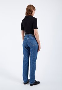 Vaqueros de mezclilla azul con un diseño de pierna recta, bolsillos traseros visibles y una etiqueta negra en la cinturilla. Combinados con una blusa negra de manga corta.