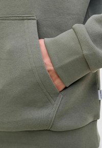 Olive green sweatshirt with ribbed cuffs, pocket detail, and smooth texture. Close-up on hand in pocket showing fabric and stitching.