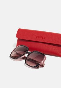 Ochelari de soare pătrați negri și roz cu lentile degradate alături de o carcasă din piele roșie embosată cu "GUESS". Accente texturate pe ramă.