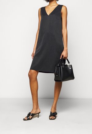 Schwarzes ärmelloses Kleid mit V-Ausschnitt, glatter Textur, knielang. Kombiniert mit schwarzen Slingback-Absätzen mit Schleife und einer strukturierten Handtasche.