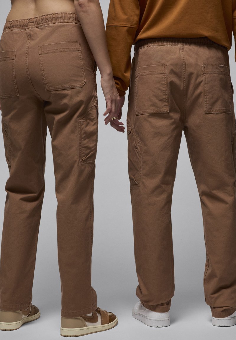 Due persone in piedi fianco a fianco, con indosso pantaloni cargo marroni abbinati, che si tengono per mano, con sneakers beige e bianche in vista.