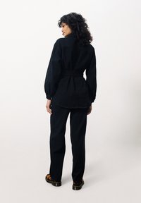 Chemise noire à ceinture avec de longues manches bouffantes, associée à un pantalon noir assorti. Le tissu semble épais avec une texture lisse. Chaussures brunes à motifs.
