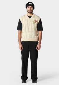 Svetr s pleteným vzorem v béžové barvě ve tvaru písmene V, zdobený květinovou výšivkou, nošený přes černé tričko, v kombinaci s černými kalhotami a obuví.