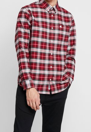 Uomo che indossa una camicia a quadri rossa, bianca e nera a maniche lunghe con pantaloni neri, in piedi con una mano in tasca.