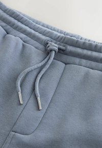 Gros plan sur la ceinture d'un pantalon de survêtement bleu clair avec une poche cousue et un cordon serré doté d'embouts argentés.