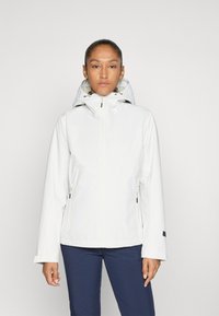 Vrouw met opgestoken haar, gekleed in een witte ritssluitingen hoodie en marineblauwe broek, staand tegen een effen lichte achtergrond.