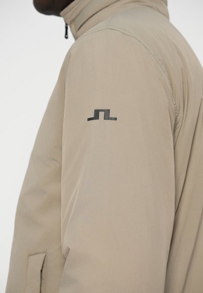 Jachetă bej cu guler înalt, închidere cu fermoar și un logo negru pe mâneca stângă. Materialul pare neted și ușor.