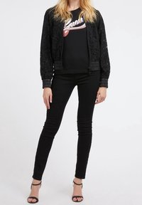 Black bomber jacket with lace detailing, paired with a black graphic t-shirt featuring a white logo, and slim black jeans. Open-toe black sandals.