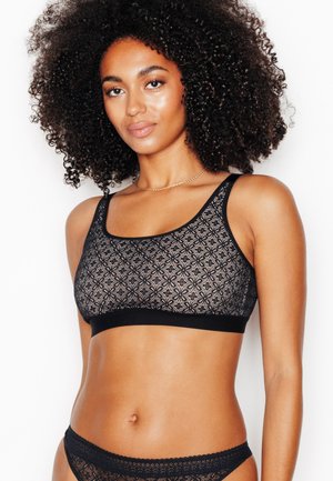 Donna con capelli ricci che indossa un bralette in pizzo nero e biancheria intima coordinata su uno sfondo bianco semplice.