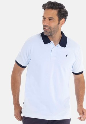 Homme portant un polo bleu clair avec un col et des bords de manches foncés, regardant légèrement sur le côté contre un fond uni.