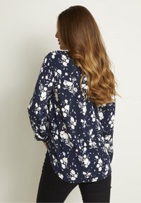 Blouse florale bleu marine avec des motifs de fleurs blanches, manches longues, coupe décontractée, ourlet incurvé et tissu texturé.