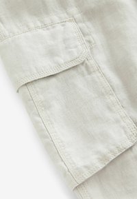 Lys beige stof med en tekstureret overflade og synlige syninger. Har en rektangulær påsyet lomme. Rent, enkelt design.