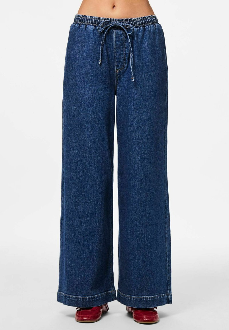 Platām kāju džinsi tumši zilā krāsā, ar elastīgu jostu un auklu. Iekļauti redzami šuvju un apakšmalu akcenti.