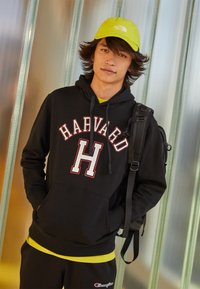Felpa nera con cappuccio con la scritta "HARVARD" e un logo "H" rosso e bianco, abbinata a una maglietta gialla e pantaloni scuri; include un cappello giallo e uno zaino.