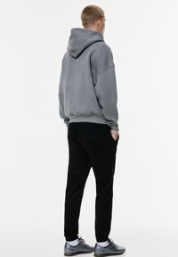Grijze hoodie met een trekkoordkap, gecombineerd met zwarte joggers en grijze sportschoenen. De stof heeft een zachte textuur en biedt een relaxte pasvorm.