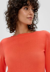 Koraal gebreide trui met een ronde hals en ribstructuur bij de kraag. De stof ziet er zacht en aansluitend uit, wat het bovenlichaam accentueert.