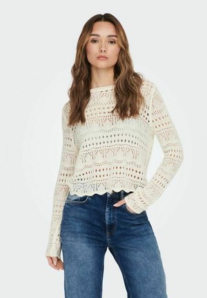 Maglione a maglia di colore crema chiaro, con motivo a traforo, orlo ondulato e maniche lunghe, abbinato a jeans di denim scuri.