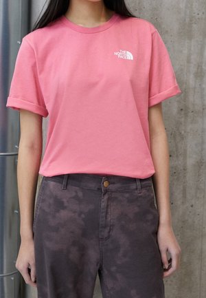 Osoba nosząca różowy t-shirt z krótkim rękawem z logo The North Face i ciemnoszare spodnie z wysokim stanem i efektem sprania na tle betonowej ściany.