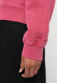 Różowa bluza z prążkowanymi mankietami, na rękawie z wytłoczonym logo. Materiał wydaje się miękki i teksturowany. Czarny tłum stanowi kontrast dla ubrania.