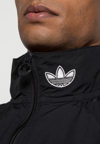 Närbild av en persons underansikte som bär en svart zip-up jacka med en vit Adidas trefoil-logotyp på kragen.