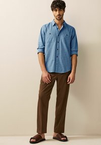 Himmelblaues Button-up Hemd mit subtilen Mustern, kombiniert mit braunen Hosen und dunklen Sandalen. Das Hemd hat zwei Brusttaschen.