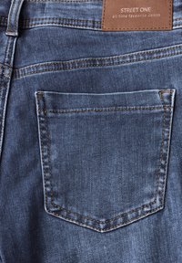 Jean bleu en denim avec une poche arrière ornée de coutures jaunes contrastantes. Étiquette en cuir à la taille avec l'inscription « STREET ONE ». 