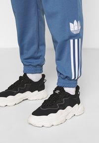 Blå sweatpants med elastiska manschetter, med vita Adidas-ränder och logotyp. Svarta sneakers med texturerad ovandel och vit sula.