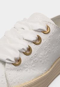 Άσπρο αθλητικό παπούτσι με υφή δαντέλας με λουλούδια, χοντρή μπεζ σόλα, χρυσά ροδέλα, και φαρδιά κορδέλα από λευκό σατέν δεμένη μέσα από τις οπές.