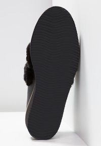 Chausson noir avec une semelle en caoutchouc texturé, des accents en fourrure douce et un dessus en matériau lisse, présentant un motif de semelle ondulé au bas.