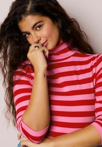 Červený a ružový pruhovaný rolák s rebrovanou textúrou; má dlhé rukávy a priliehavý strih. Strieborný prsteň na ruke.